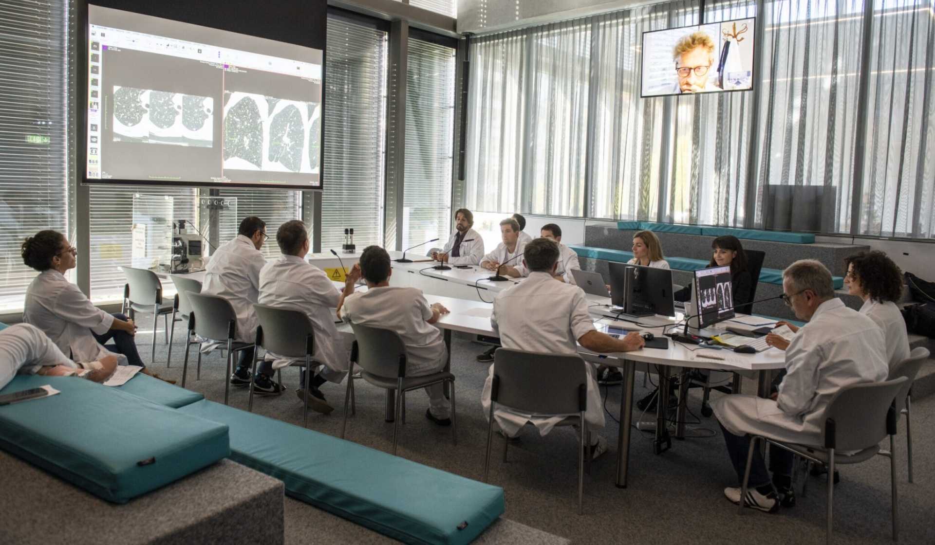 Réunion médicale dans une salle moderne : des professionnels en blouse blanche analysent des images radiologiques projetées à l’écran et collaborent autour de plusieurs ordinateurs.